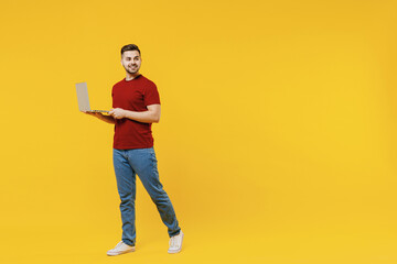 Full length satisfied smiling young man in red t-shirt casual clothes using laptop pc computer work look aside isolated on plain yellow color wall background studio portrait. People lifestyle concept.