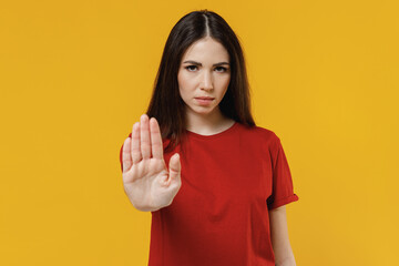 Frowning serious confident beautiful young brunette woman 20s wears basic red t-shirt showing stop gesture with palm isolated on yellow background studio portrait. People emotions lifestyle concept.