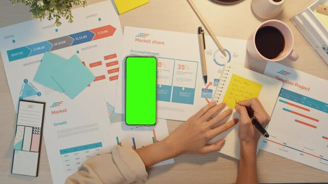 Top view of young Asia lady freelance use phone with blank green screen mock up display for advertising text and write worksheet finance graph account chart market plan at office night. Chroma key.