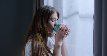 Smiling Caucasian woman standing by window with cup of tea. Young woman drinking hot coffee looking outside at landscape.
