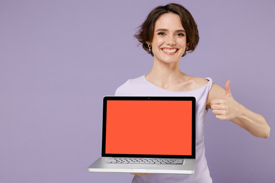 Young smiling freelancer woman in white t-shirt hold use work on laptop pc computer with blank screen workspace area show thumb up like gesture isolated on pastel purple background studio portrait