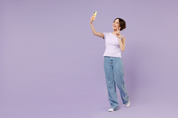 Full length young smiling fun woman in white t-shirt do selfie shot on mobile cell phone post photo on social network show victory v-sign gesture isolated on pastel purple background studio portrait