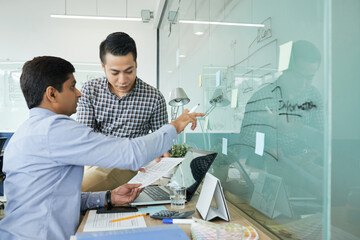 UX and UI designers working on project together, they are discussing chart on glass wall and documents with interface layouts
