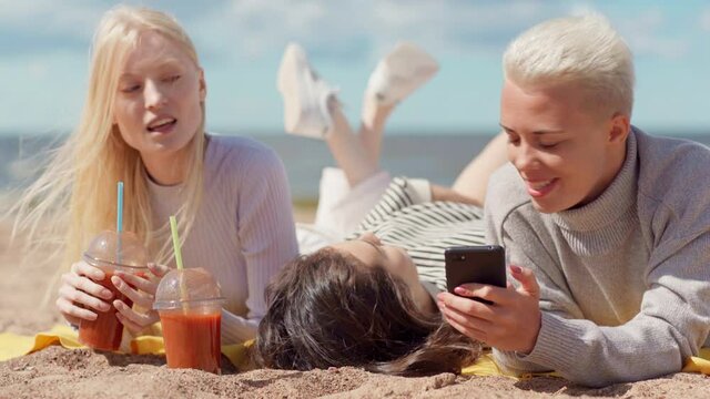 Group of three relaxed girlfriends with smoothie drinks lying down on beach and discussing photos or messages in social media looking on cell phone screen. Young women spending vacation on sea coast