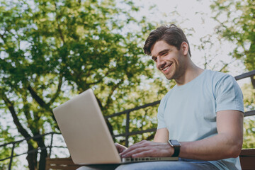 Young freelancer smiling happy satisfied student man in blue t-shirt sit on bench work with laptop pc computer rest relax in sunshine spring green city park outdoors on nature. Urban leisure concept.