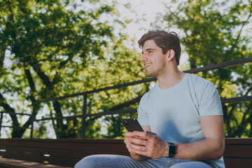 Minded young happy smiling man 20s wear blue t-shirt sit on bench using mobile cell phone chat online look aside rest relax in sunshine spring green city park outdoor on nature Urban leisure concept.