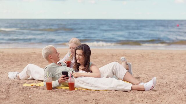 Group of three happy relaxed girlfriends with smoothie drinks lying on sea beach together and discussing photos, news or text messages in social media looking at smartphone screen