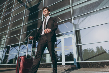 Bottom view pensive young traveler businessman man 20s in black dinner suit walk go stand outside at international airport terminal with suitcase valise looking aside. Air flight business trip concept