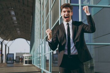 Happy young traveler businessman man 20s in black classic tie suit standing outside at international airport terminal do winner gesture clench fist People air flight business trip lifestyle concept.