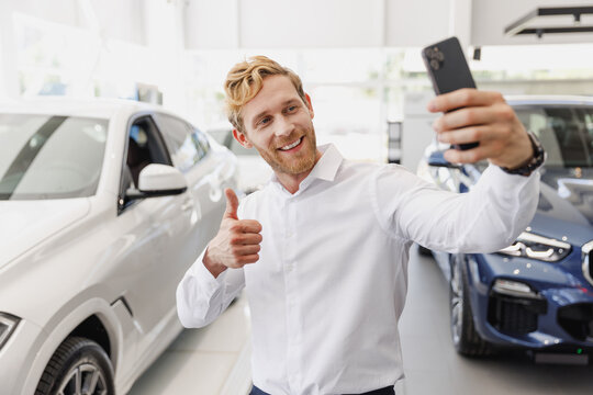 Man customer buyer client in white shirt do selfie shot on mobile cell phone show thumb up choose auto want buy new car automobile in showroom vehicle dealership store motor show indoor Sales concept.