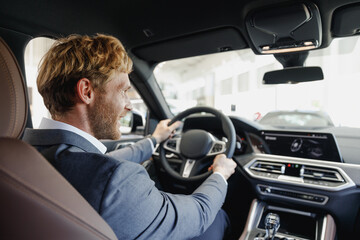 Back view smiling man customer buyer client in classic grey suit driving car hold wheel choose auto...