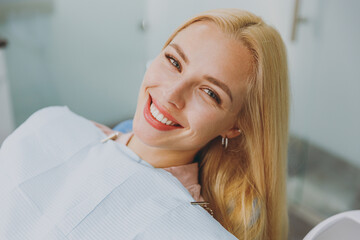 Close up fun young smiling happy calm woman covered by napkin sitting at dental office chair indoor...