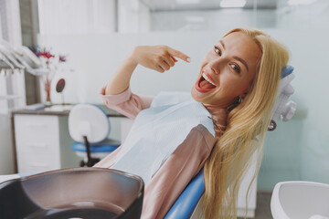Obraz na płótnie Canvas Young smiling happy woman covered by napkin point index finger on healthy toothy smile sit at dental office chair indoor light modern cabinet wait for stomatologist Healthcare oral enamel treatment