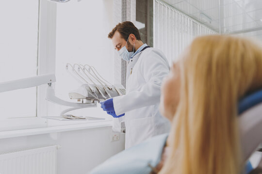Young man doctor in white gown prepare for procedure patient blonde woman sit at dentist office chair indoor cabinet near stomatologist wait for oral examination Healthcare caries enamel treatment.