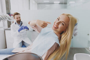 Young smiling happy woman 20s with dental napkin point finger on mouth sit at dentist office chair...