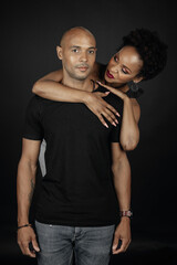 latin man and woman posing on black background, girl looks at him and smiles; he looks at you.