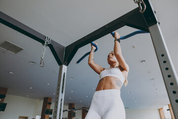 Bottom view young skinny caucasian strong sporty athletic sportswoman woman 20s wear white sportswear warm up training pulls up on horizontal bar on in gym indoors Workout sport motivation concept.