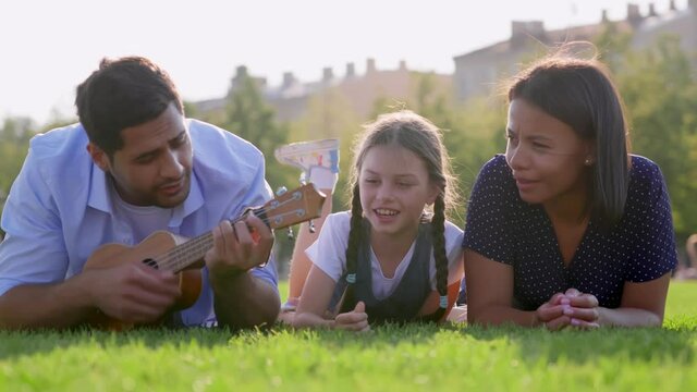 Portrait of multiethnic family lying on green lawn in park playing ukulele