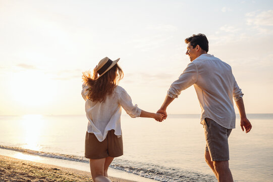 Back rear view young happy couple two friends family man woman in white shirt clothes hold hands walk run stroll together at sunrise over sea beach ocean outdoor exotic seaside in summer day evening.
