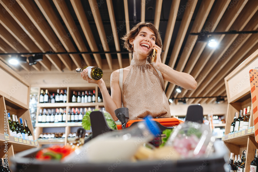 Wall mural Young smiling happy woman in casual clothes shopping at supermaket store with grocery cart hold white wine alcohol hold bottle talk speak mobile phone inside hypermarket Purchasing gastronomy concept