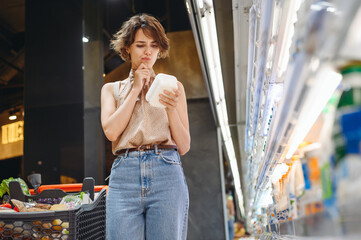 Young minded woman 20s in casual clothes backpack shopping at supermaket store buy dairy produce take hold container read shelf life prop up chin inside hypermarket Purchase gastronomy food concept.