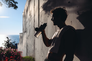 Side view young serious sportive strong caucasian sunlit sportsman man with naked torso training hold in hand water bottle on grey wall background near sea resort Outdoor courtyard sports concept.