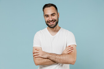 Young fun smiling happy man 20s wearing casual white t-shirt looking camera hold hands crossed folded isolated on plain pastel light blue color background studio portrait. People lifestyle concept