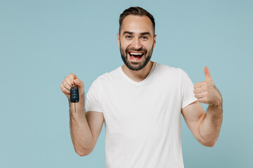 Young smiling happy fun man 20s in blank print design white t-shirt hold in hand car key fob keyless system show thumb up gesture isolated on plain pastel light blue color background studio portrait