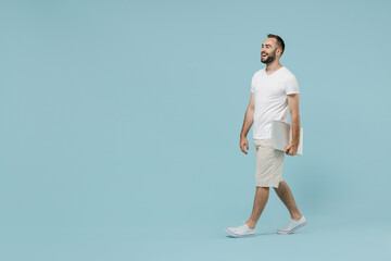 Fototapeta na wymiar Full length side view young confident smiling happy minded man 20s in white t-shirt walking going hold closed laptop pc computer look aside isolated on plain pastel light blue color background studio