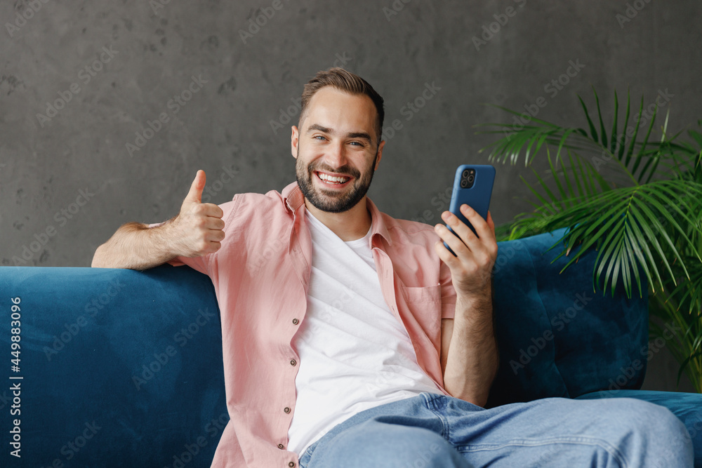 Wall mural young smiling happy man in casual clothes hold in hand use mobile cell phone show thumb up sitting o