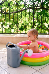Child sits in an inflatable mini-pool, next to a garden watering can