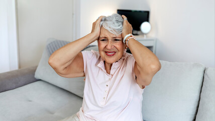 Tired older woman sitting on comfortable sofa in living room, touching forehead. Exhausted mature lady suffering from head ache at home. Unhappy elderly grandmother worrying about bad news alone.