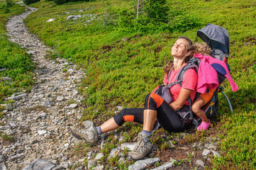 Naklejka na ściany i meble A mother with her sleeping daughter in a carrier backpack is resting on the grass before climbing a stone path in the Carpathian Gorgan. Ukraine