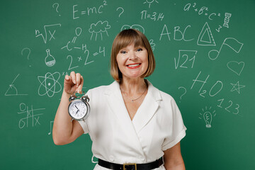 Smiling intelligent teacher mature elderly lady woman 55 wear white shirt hold in hands clock isolated on green wall chalk blackboard background studio. Education in high school September 1 concept.