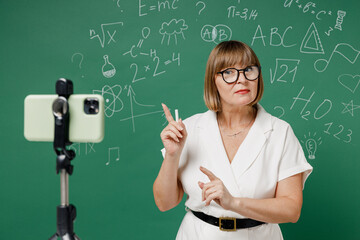 Smart serious teacher mature elderly lady woman 55 wear shirt glasses work with mobile cell phone on tripod on quarantine explain new material isolated green wall chalk blackboard background studio.
