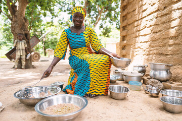 Malian housewife sourrounded by plates with food, pots and kitchen utensils cooking a meal for her...