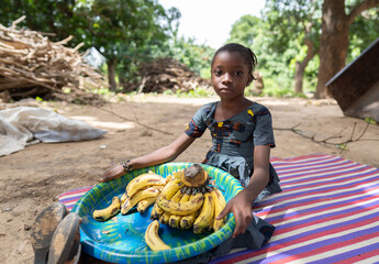 Sad looking small black African girl sitting on a mat holding a big plate of bananas, waiting for...