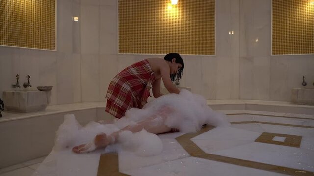 The traditional oriental massage uses a lot of foam. The woman lies on her stomach and is completely covered in foam. Turkish woman massages her back. Selective focus. Step 14