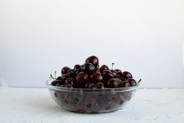 A transparent bowl full of sweet cherries on a rough table against a white wall.