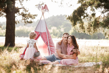 Mom, dad and little daughter are sitting next to wigwam decoration in the park. Family spending time outdoor in summer, having fun together. Girl are dressed in pink dress © Andriy Medvediuk