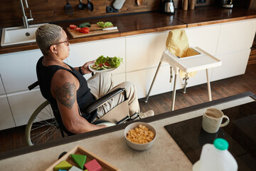 High angle portrait of contemporary tattooed woman with disability in kitchen, baby chair in shot, copy space
