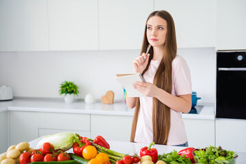 Portrait of attractive minded long-haired girl writing to-do list cooking vegan dish in light white home kitchen indoors