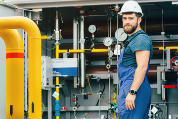 Portrait of a gas worker in a helmet and a working overalls against the background of equipment with pipes and pressure gauges. Gas odorization unit. Gas distribution station operator.