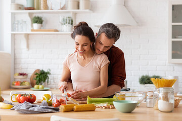 Happy wife and husband cooking romantic dinner at home kitchen. Man hugging woman from back. Couple chopping vegetable together. Fresh salad recipe preparation. Loving relationship. Family portrait