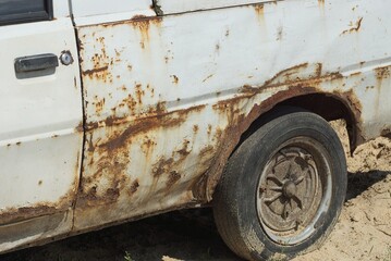 part of an old white passenger car in brown rust with a black wheel on gray sand in the street