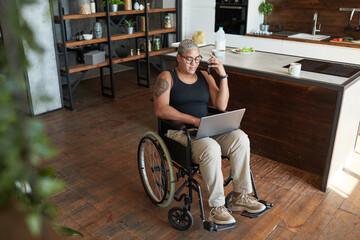 Full length portrait of contemporary tattooed woman with disability using laptop while working from...