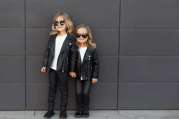 Street style portrait of two cute pretty sisters 2-3 years old in same outfits: white t-shirts, leather black jackets and leggings. Best friends together outdoors.