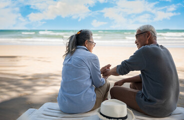 Romantic of Asian senior couple tourist is sitting hand in hand and talking on beach chair with hat in summer vacation.