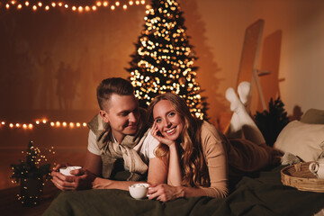 Naklejka na ściany i meble The concept of Christmas. A happy man and a woman in love celebrate the winter New Year holidays at night in a cozy house. A couple in love are kissing hugging together watching a movie eating popcorn