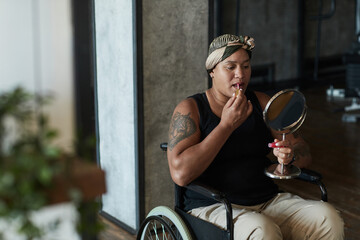 Portrait of African-American woman in wheelchair putting on lipstick while holding mirror in home interior